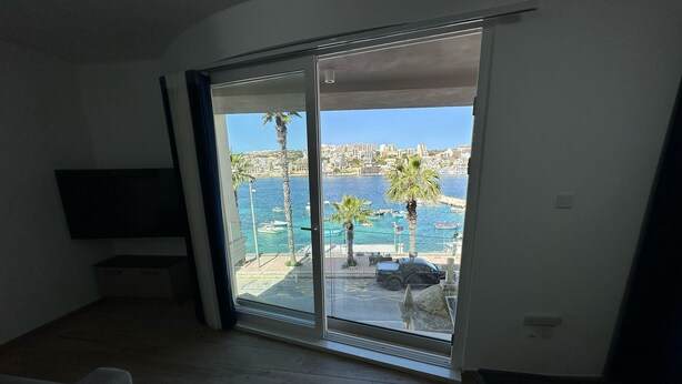 A room with a view of a coastal area, featuring a sliding glass door leading to an outdoor space with palm trees and a body of water.