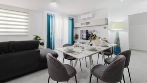 Uma sala de estar moderna com um sofá cinzento, uma mesa de jantar posta para quatro pessoas e uma TV na parede.