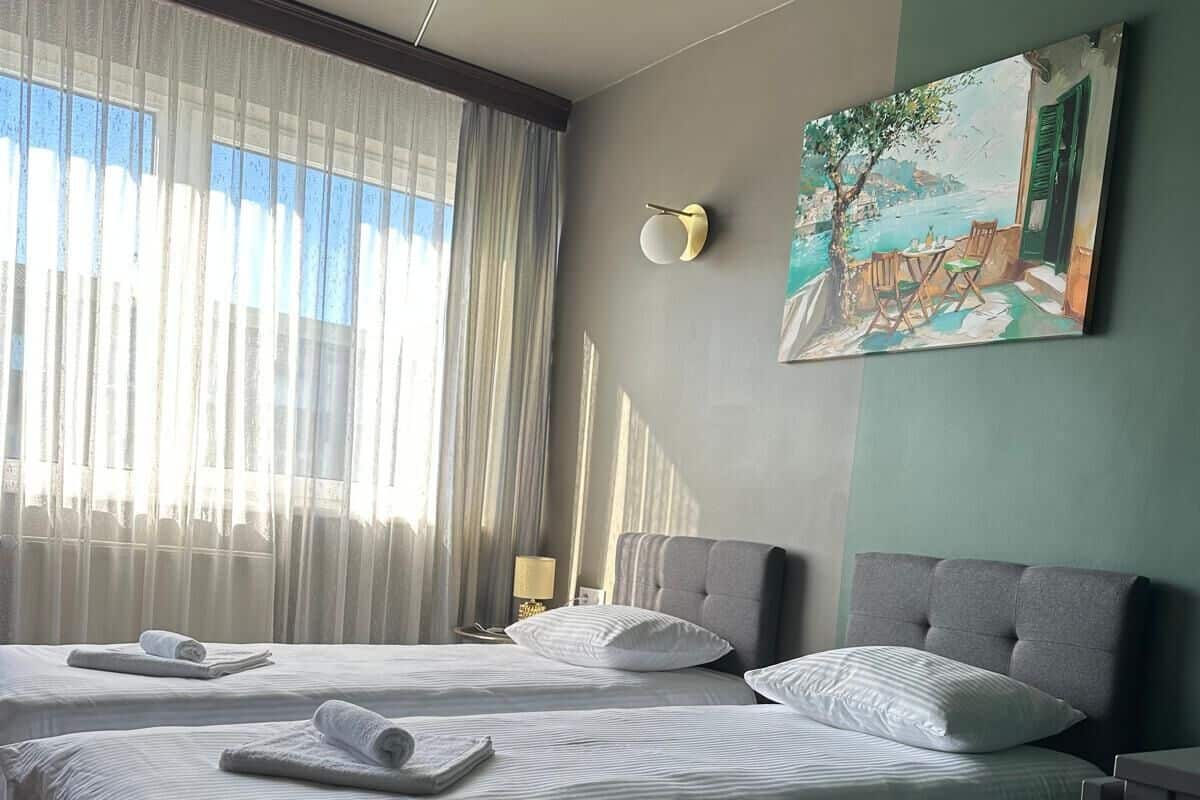 A hotel room with two beds, a painting on the wall, and a window with sheer curtains.