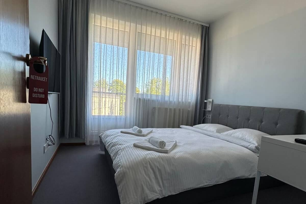 A modern hotel room with a large bed, a bedside table, a TV mounted on the wall, and a window with curtains.