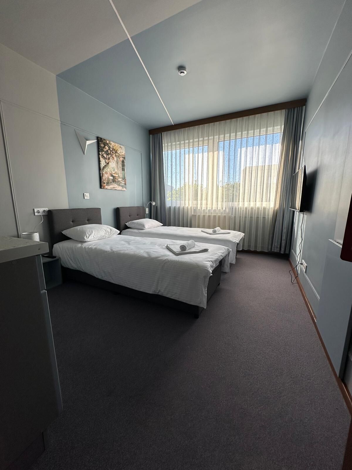 A hotel room with two beds, a flat-screen TV, a painting on the wall, and a window with sheer curtains.