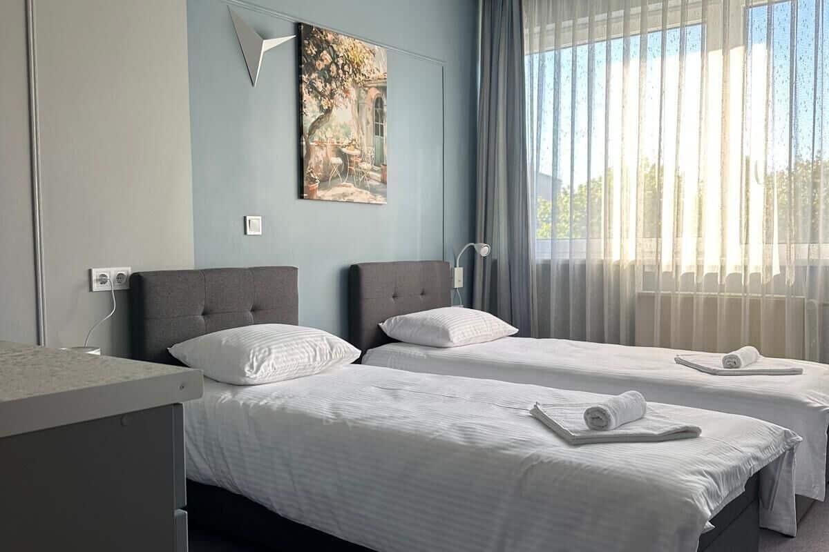 A hotel room with two beds, a large window with sheer curtains, a painting on the wall, and a modern design.