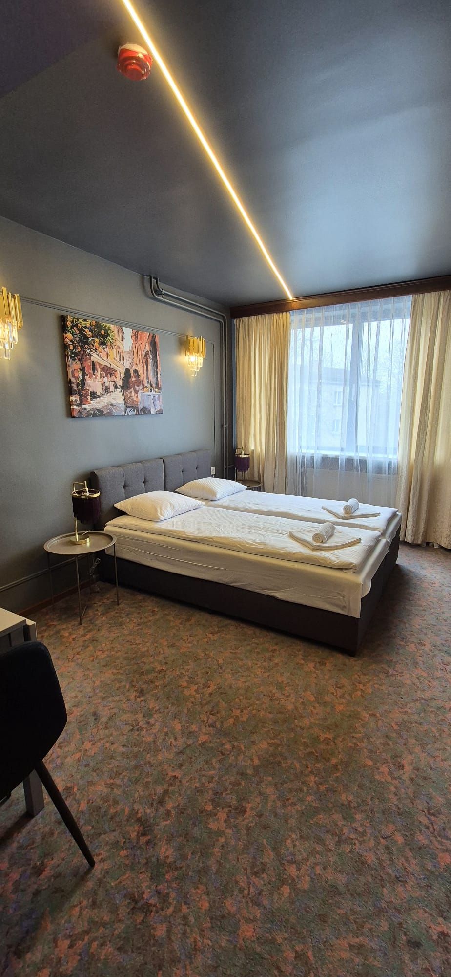 Habitación de hotel con una cama grande, una mesita redonda pequeña, una silla, un cuadro en la pared y una ventana con cortinas.