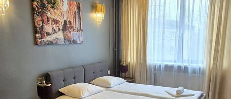 Habitación de hotel con una cama grande, una mesita redonda pequeña, una silla, un cuadro en la pared y una ventana con cortinas.