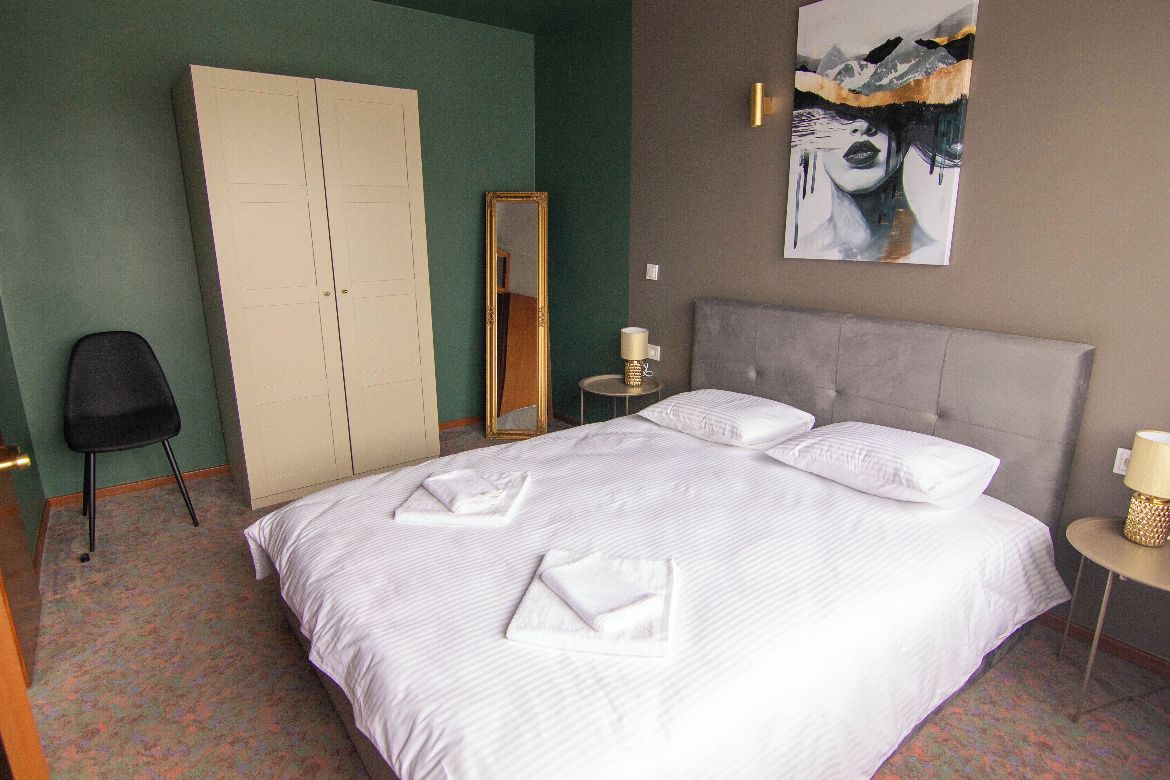 A neatly made bed with white linens and towels, a grey headboard, a bedside table with a lamp, a full-length mirror, and a wardrobe.