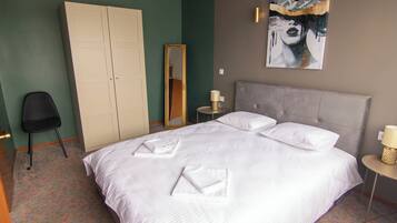 A neatly made bed with white linens and towels, a grey headboard, a bedside table with a lamp, a full-length mirror, and a wardrobe.