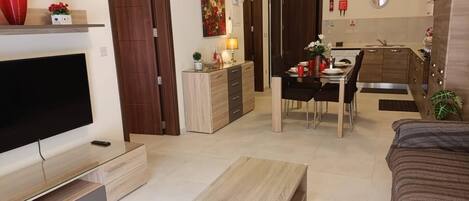 A modern living room with a flat-screen TV, a wooden coffee table, and a sofa. There is a dining area with a table and chairs, and a kitchen with wooden cabinets and a stove.
