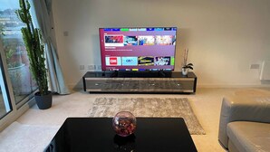 A modern living room with a flat-screen TV, a glass coffee table, a grey sofa, and a potted plant. - The Penthouse in Norfolk (Norwich)