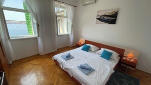 A hotel room with a large bed, two bedside tables with lamps, a view of the sea through the windows, and an air conditioning unit on the wall. - Apartment Perovic (Jadranovo)