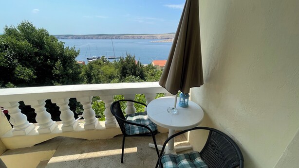 A balcony with a white balustrade, two wicker chairs with checkered cushions, a small round table, and a brown umbrella. - Apartment Perovic (Jadranovo)