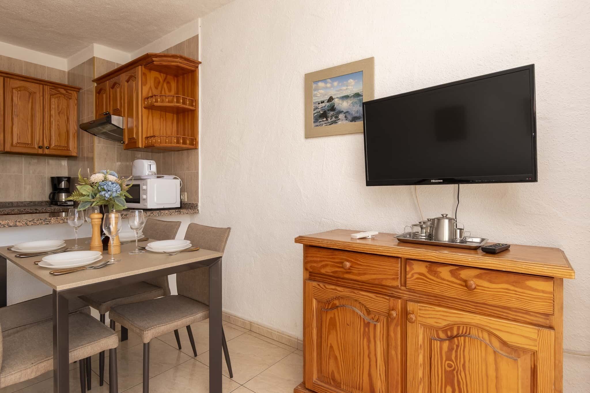 A modern kitchen with wooden cabinets, a dining table set for four, a flat-screen TV mounted on the wall, and a coffee maker.