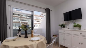 A dining area with a table set for two, a white cabinet, a TV mounted on the wall, and a balcony with a view of buildings. - Las Floritas 31 - One Bed (Playa de las Américas)