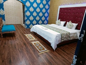 Quarto com chão de madeira, cama com roupa de cama branca e cabeceira vermelha, parede com padrão azul e cadeira de madeira com almofada azul.