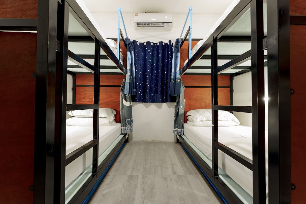 Un couloir étroit, avec des lits superposés de chaque côté, un rideau orné de motifs en forme d’étoiles et un appareil de climatisation fixé au mur.