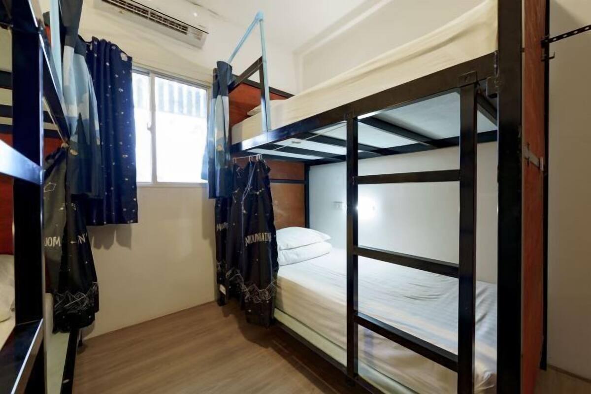 Une chambre avec un lit superposé, une fenêtre, un rideau et un porte-manteau sur lequel sont suspendus des vêtements.
