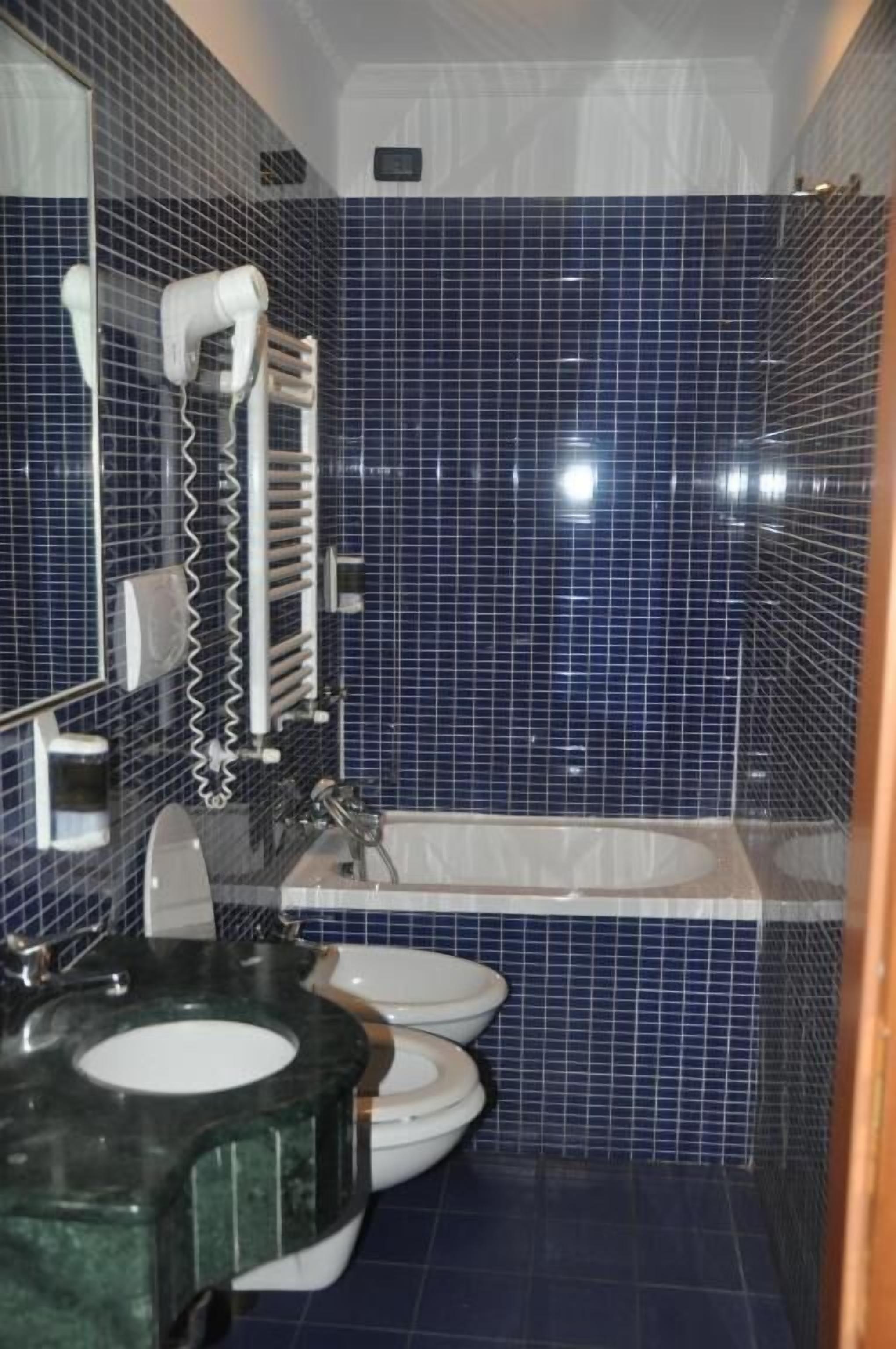 A bathroom with blue tiles, a sink, two toilets, and a bathtub.