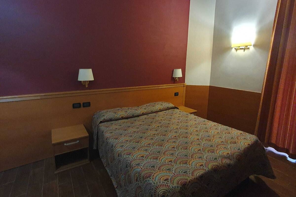 A hotel room with a bed, two bedside tables, a nightstand, and a lamp on the wall.