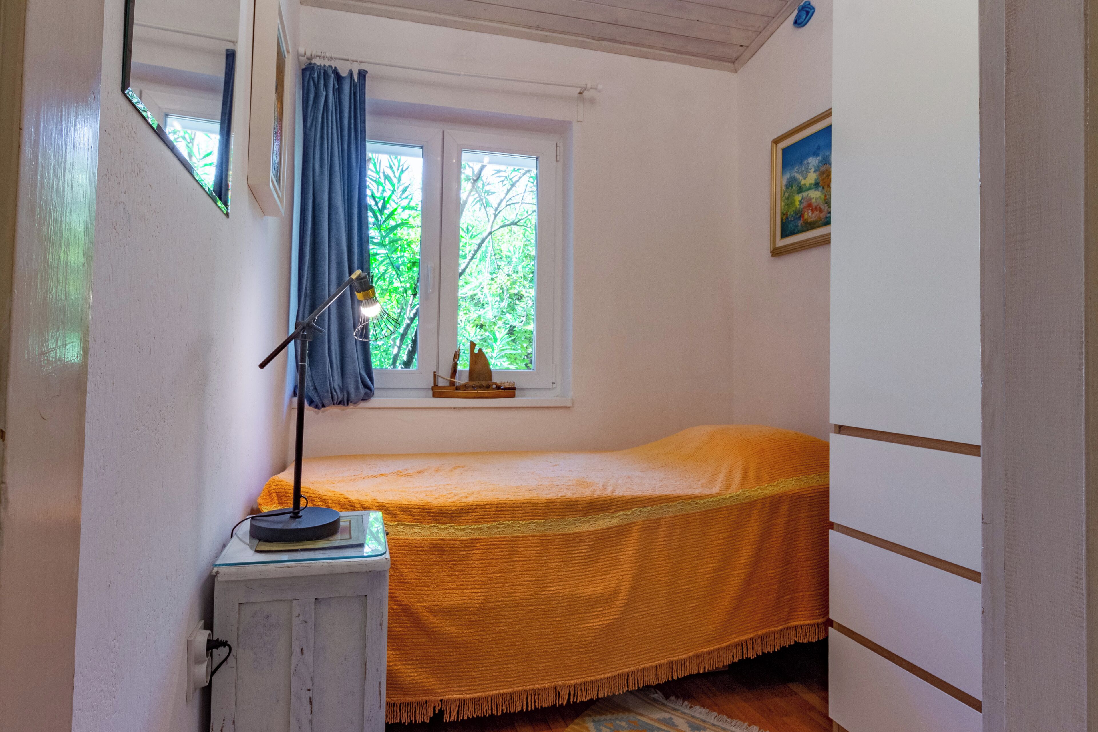 A small bedroom with a single bed, a desk with a lamp, a window with curtains, and a painting on the wall.