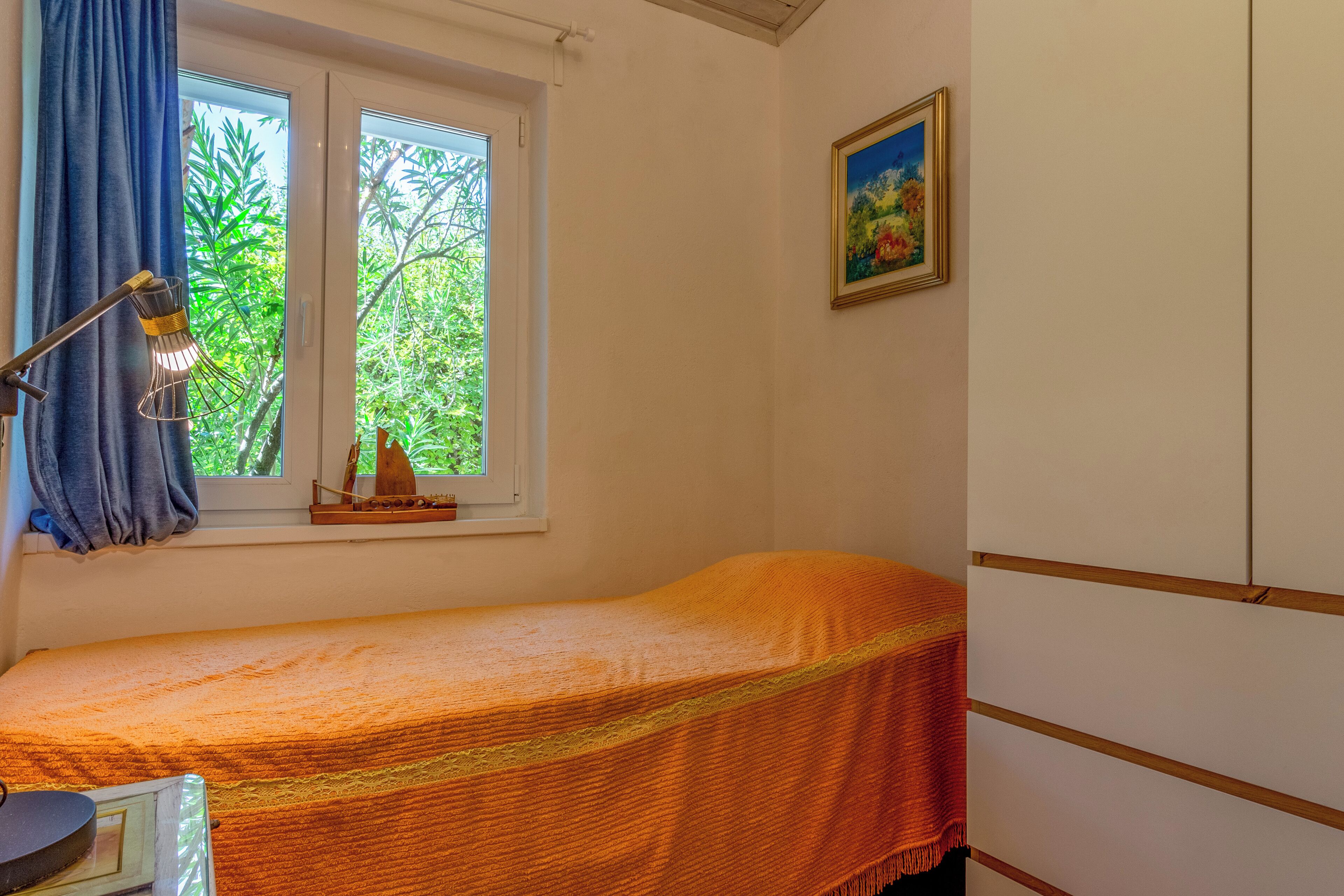 A small, single-bed room with a window, a desk, and a framed picture on the wall.