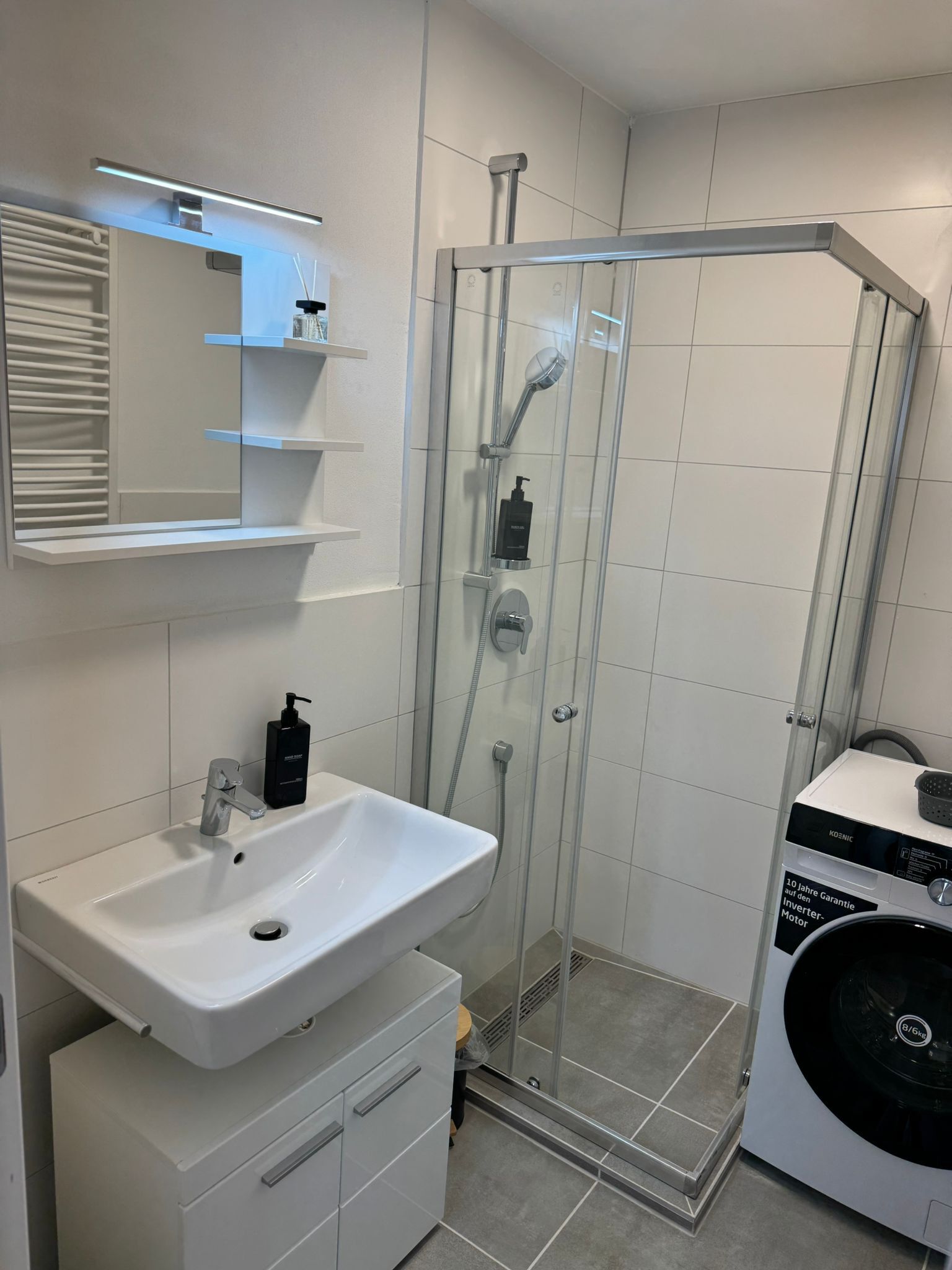 A modern bathroom with a glass-enclosed shower, a white sink with a soap dispenser, and a built-in washing machine.