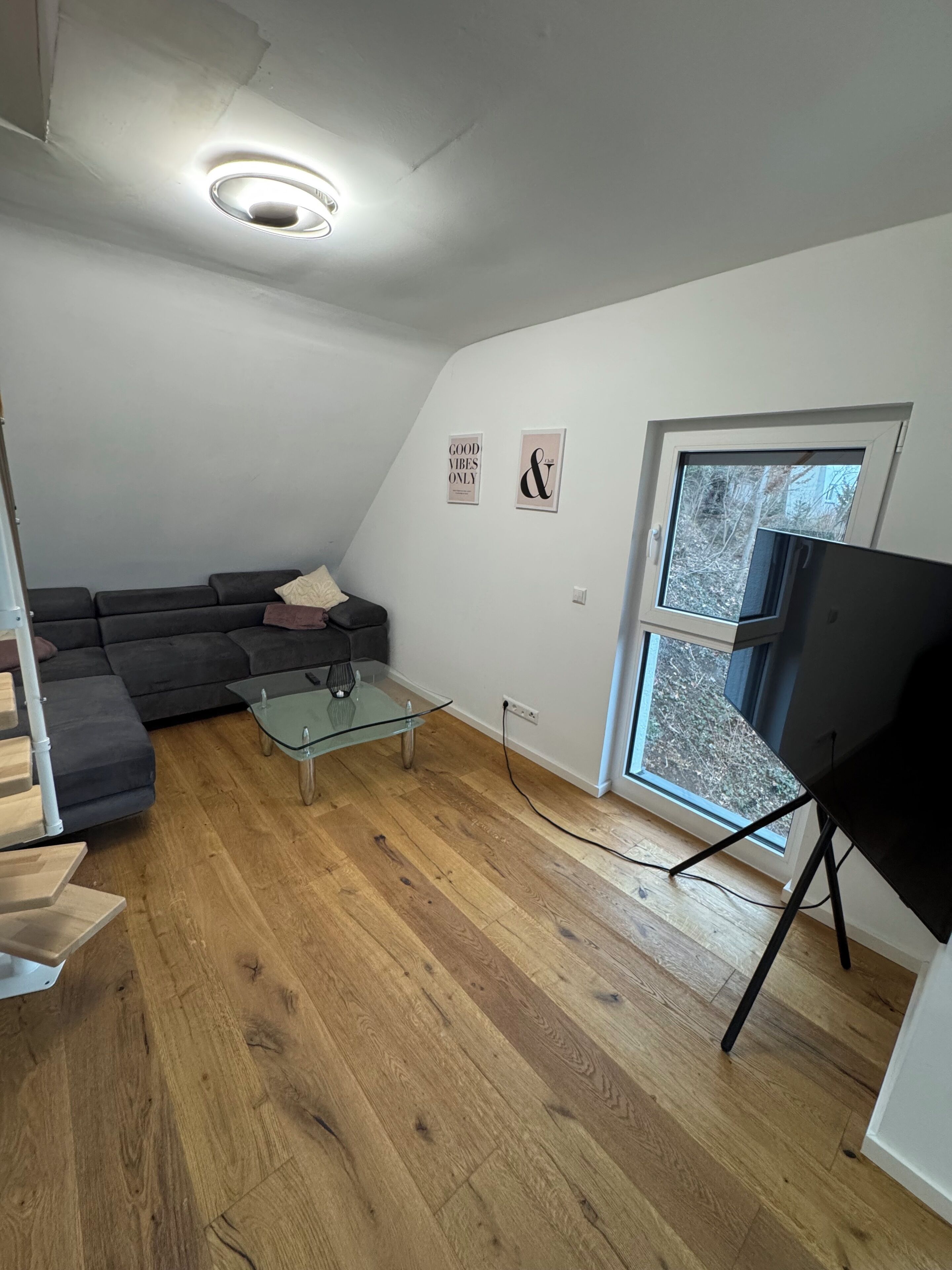 A modern living room with a sectional sofa, a glass coffee table, a flat-screen TV on a stand, and a window with a view of trees.
