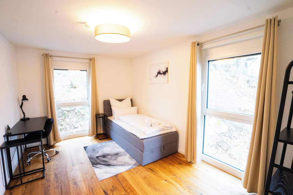 Une chambre à coucher avec un lit, un bureau et une chaise. De grandes fenêtres sont présentes, et elles sont dotées de rideaux.