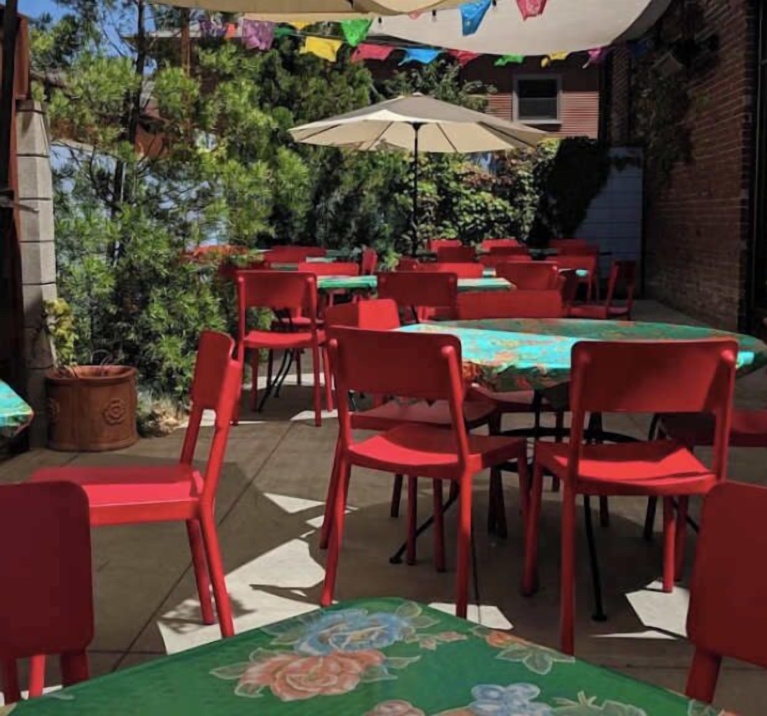 Un patio al aire libre con sillas y mesas rojas, rodeado de plantas en macetas y un muro de ladrillo.
