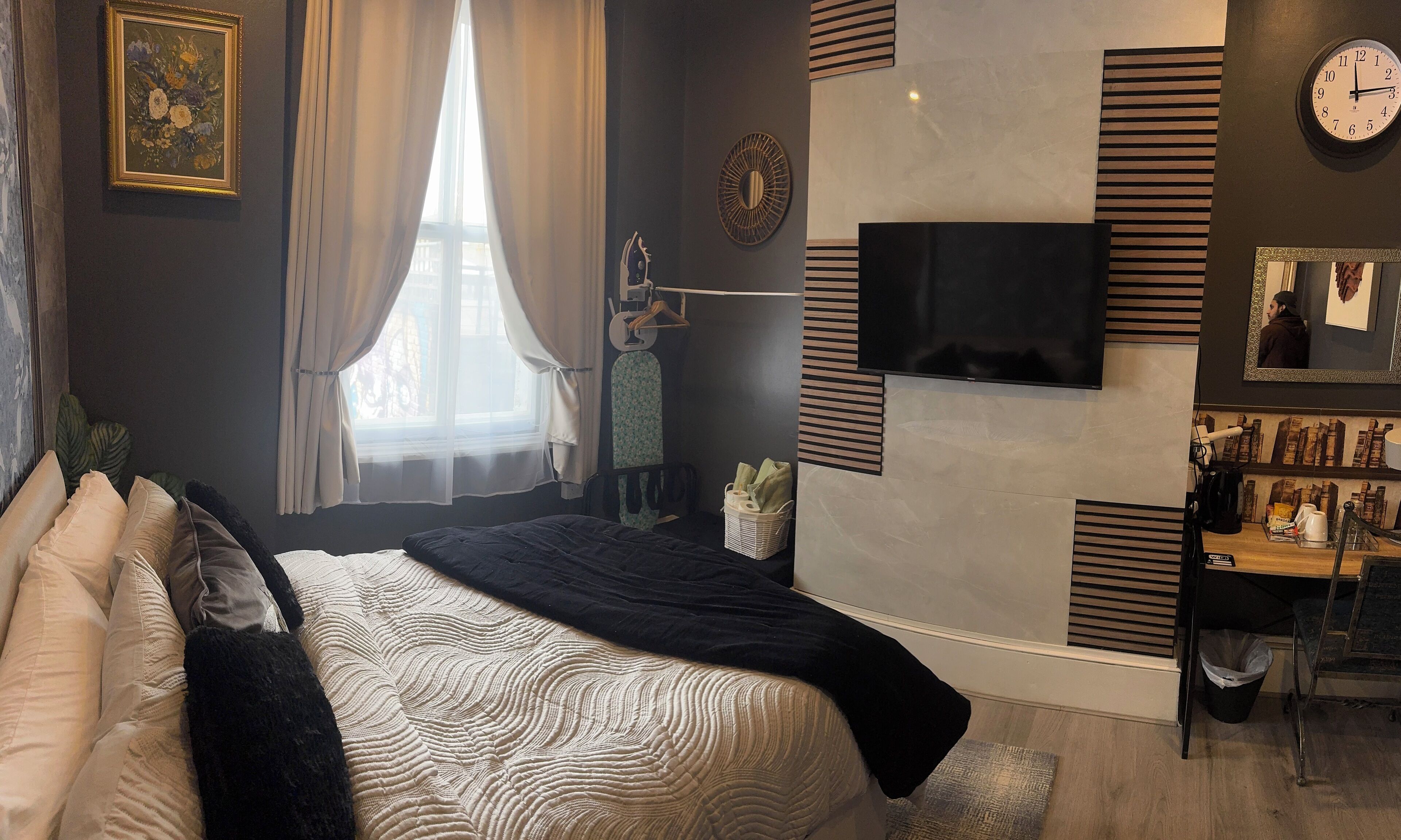 A modern bedroom with a large bed, a flat-screen TV mounted on the wall, a window with sheer curtains, and a coffee machine on a wooden counter.