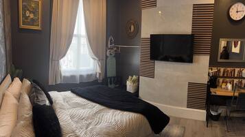 A modern bedroom with a large bed, a flat-screen TV mounted on the wall, a window with sheer curtains, and a coffee machine on a wooden counter.