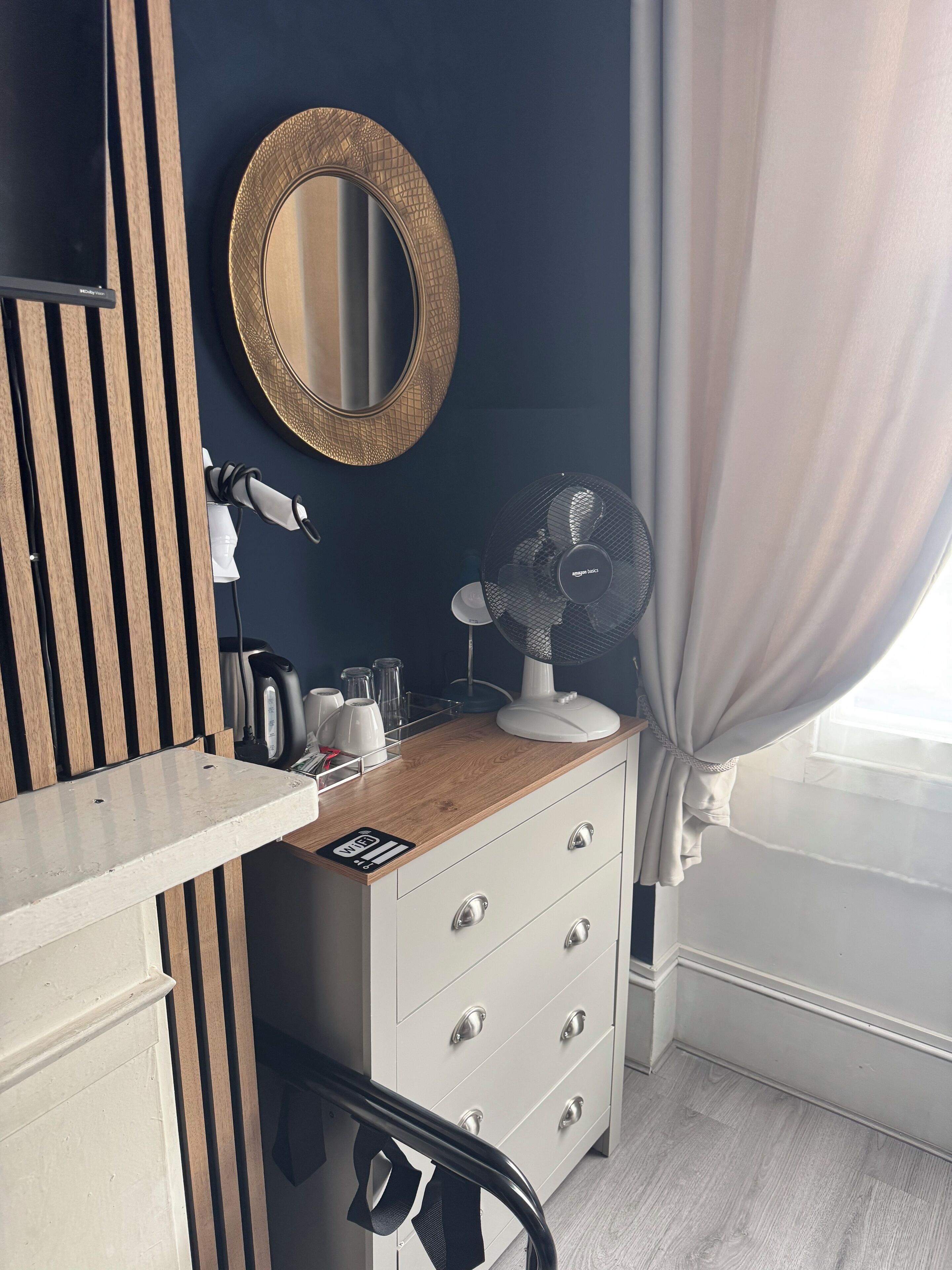 Une chambre d’hôtel avec une coiffeuse en bois, un miroir, une commode blanche à tiroirs, un ventilateur sur pied et une fenêtre avec des rideaux.
