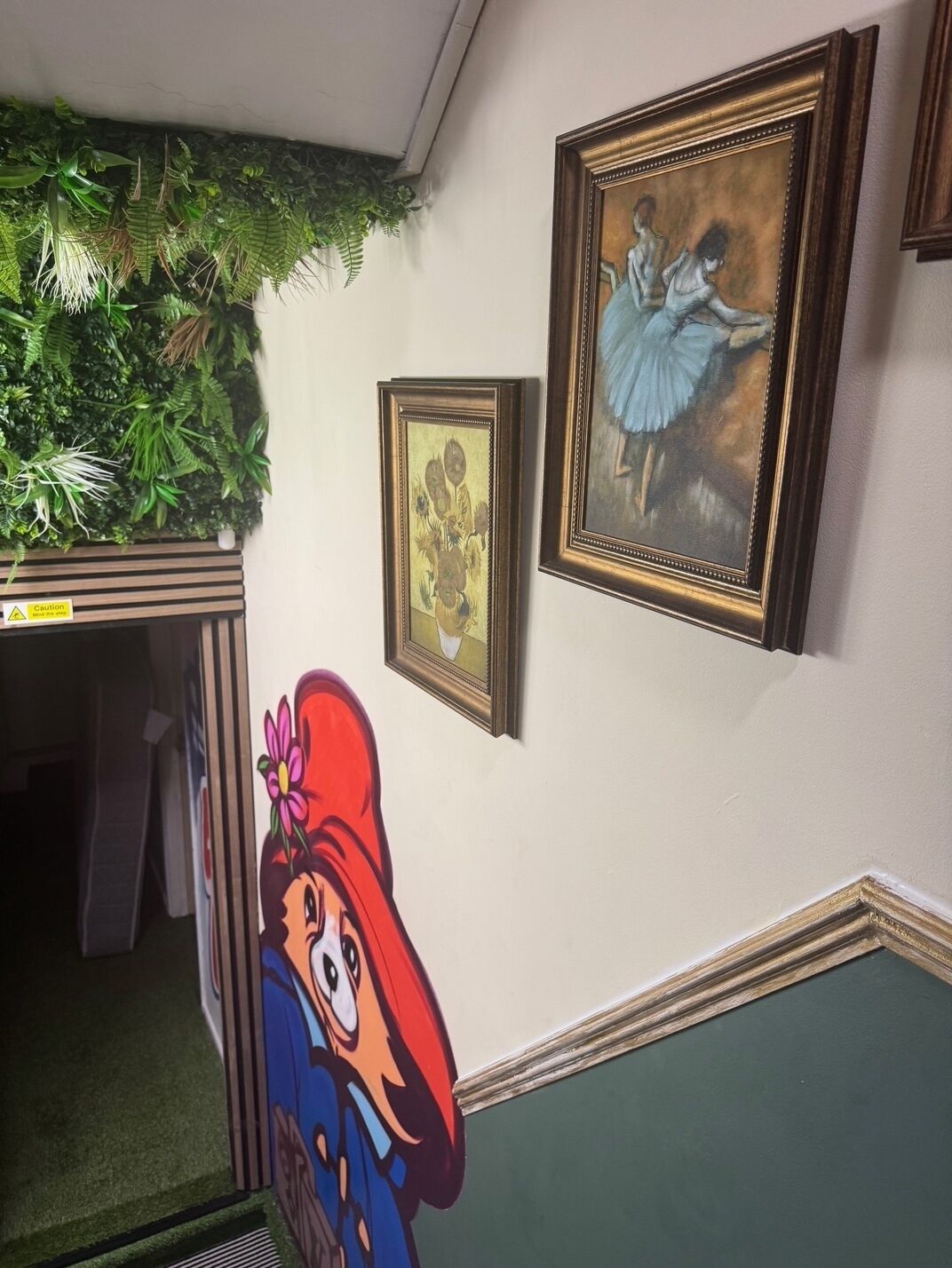 Un pasillo con pared verde, un cuadro enmarcado y un recorte de un personaje de caricatura.
