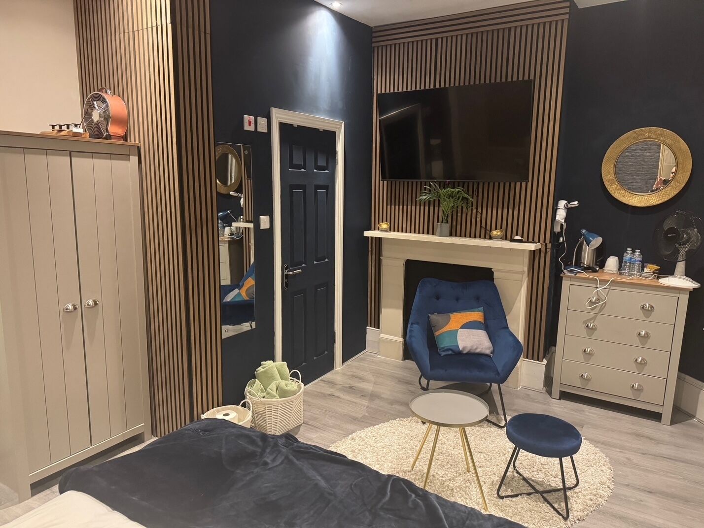 Een moderne slaapkamer met een bed, een blauwe fauteuil, een klein rond tafeltje, een dressoir, een spiegel en een open haard.