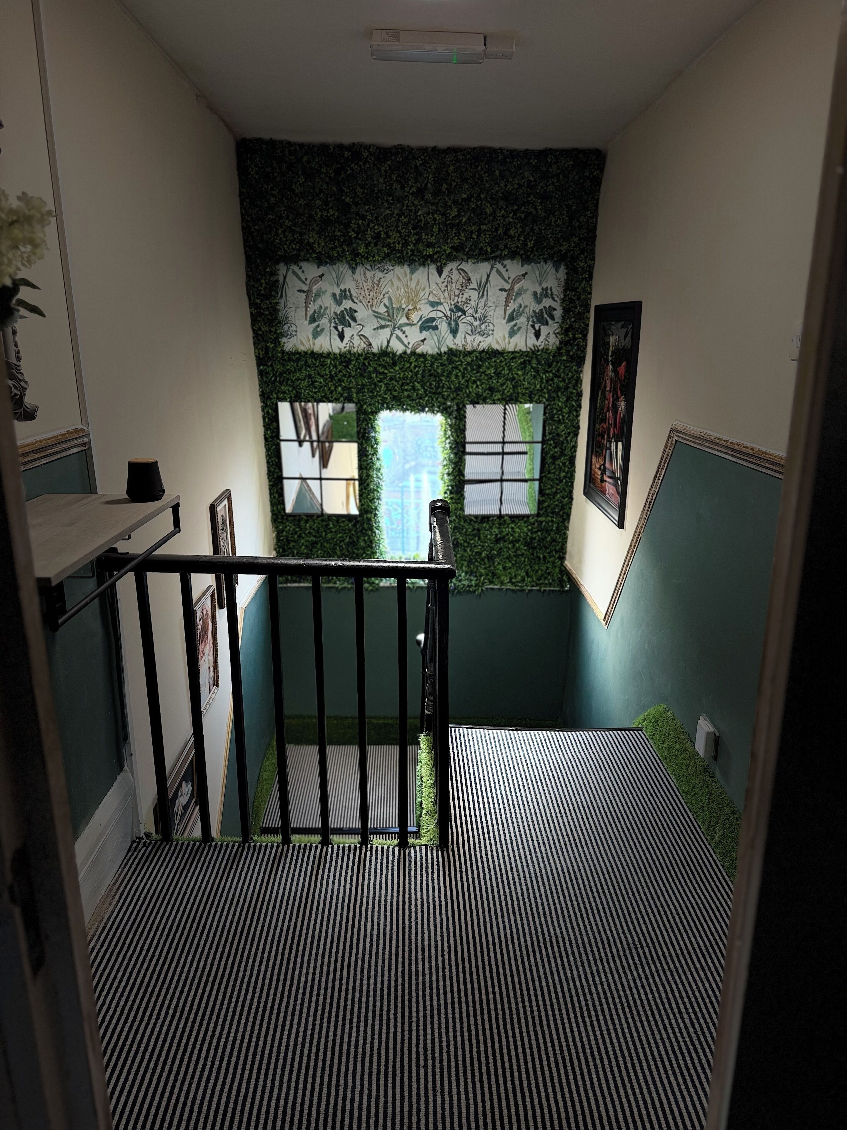 A staircase with a black railing leading to a landing. The landing has a patterned floor and a green wall with a window.