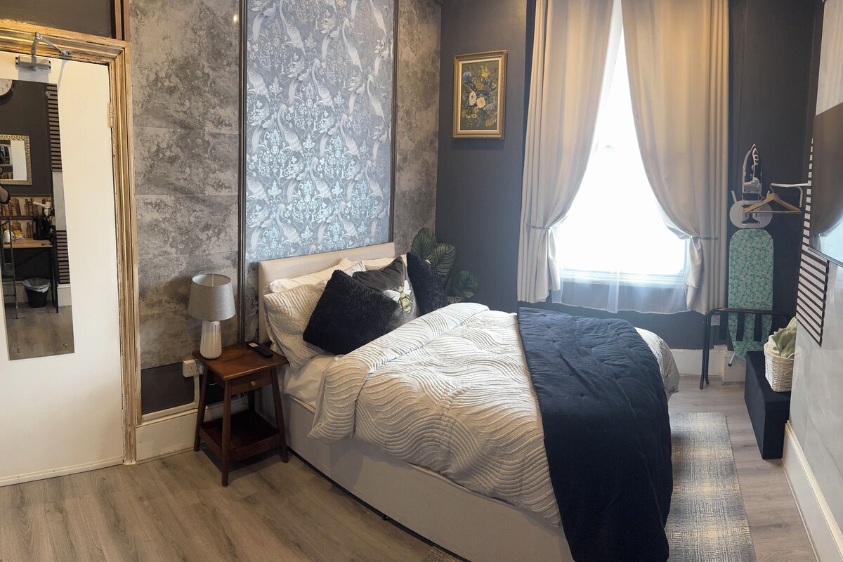A bedroom with a bed, a nightstand, a lamp, a mirror, a TV, and a window with curtains.