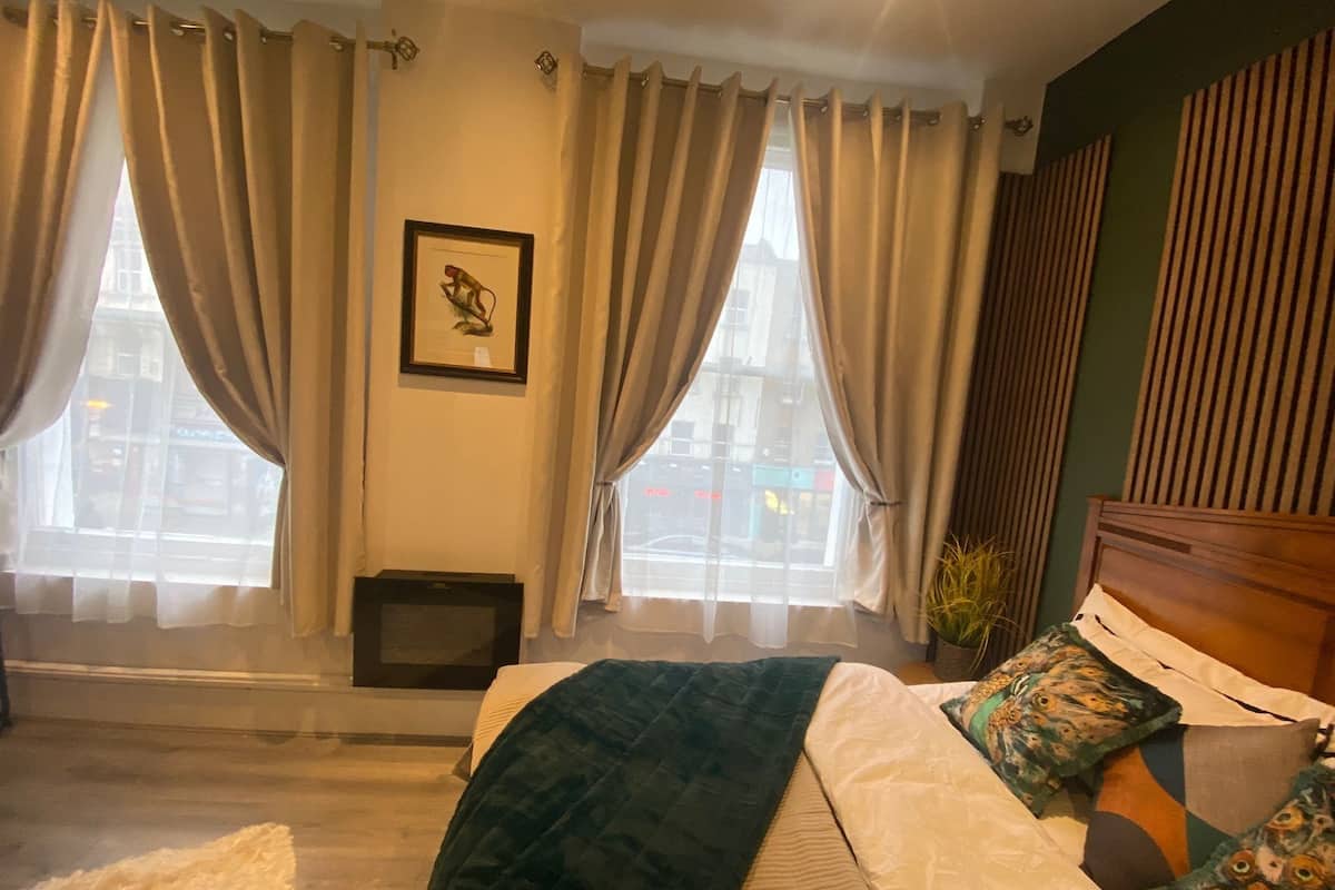Une chambre à coucher avec un lit, des rideaux, un tableau encadré et une fenêtre donnant sur des immeubles.