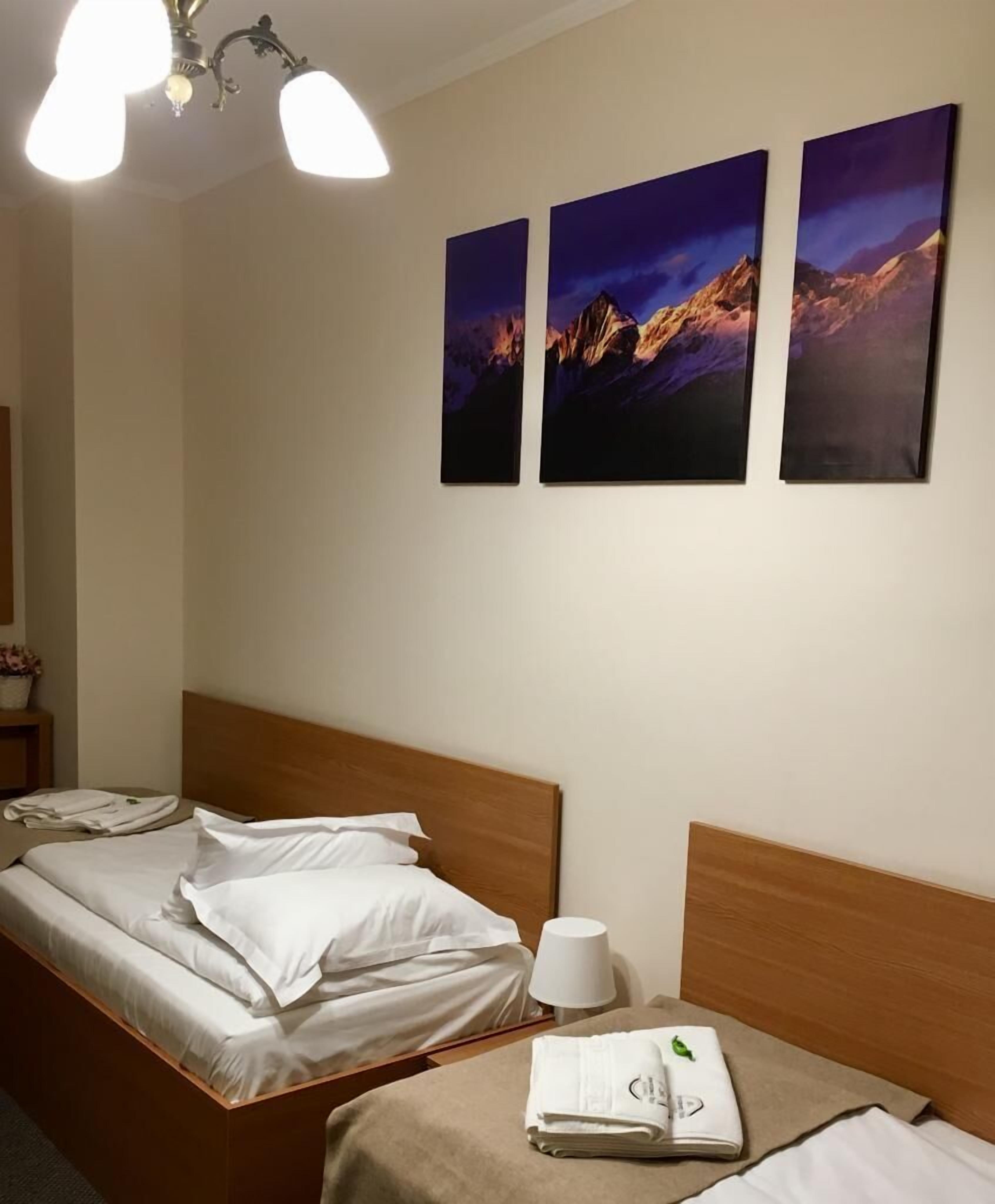 A hotel room with a bed, a bedside table with a lamp, and three paintings on the wall.