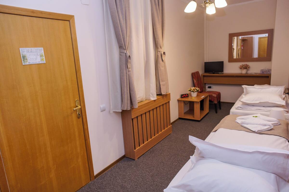 Habitació d'hotel amb una porta de fusta, un llit amb roba de llit blanca, una petita taula de nit amb una cadira, una televisió i un mirall a la paret.