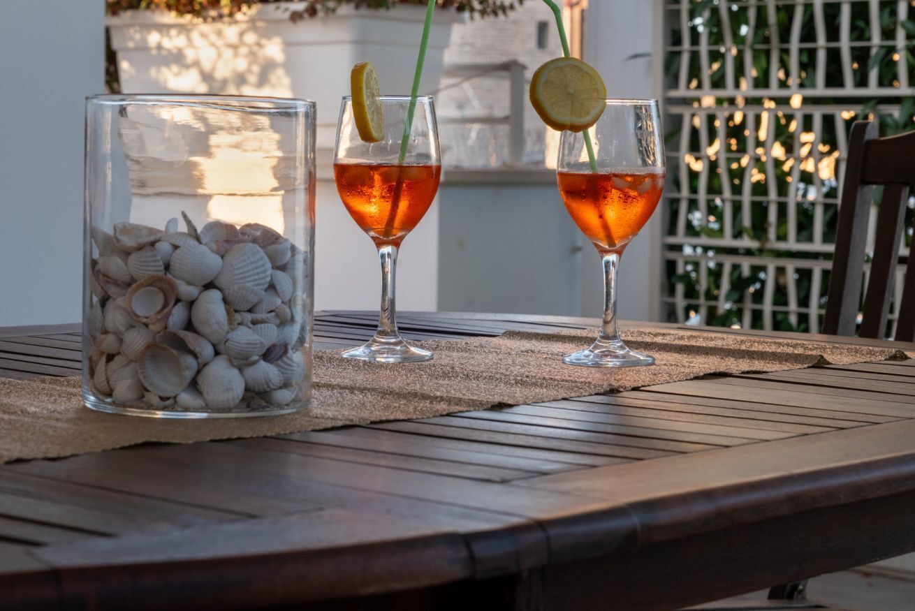 A table with two glasses of amber-colored drink, each garnished with a lemon slice and a straw. A clear glass jar filled with seashells is placed on the table.