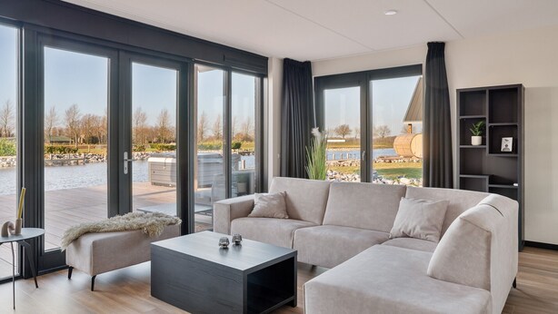 Una sala de estar moderna con un sofá grande, una mesa de centro y una estantería. Cuenta con ventanas amplias con vistas a un lago y mobiliario de exterior.