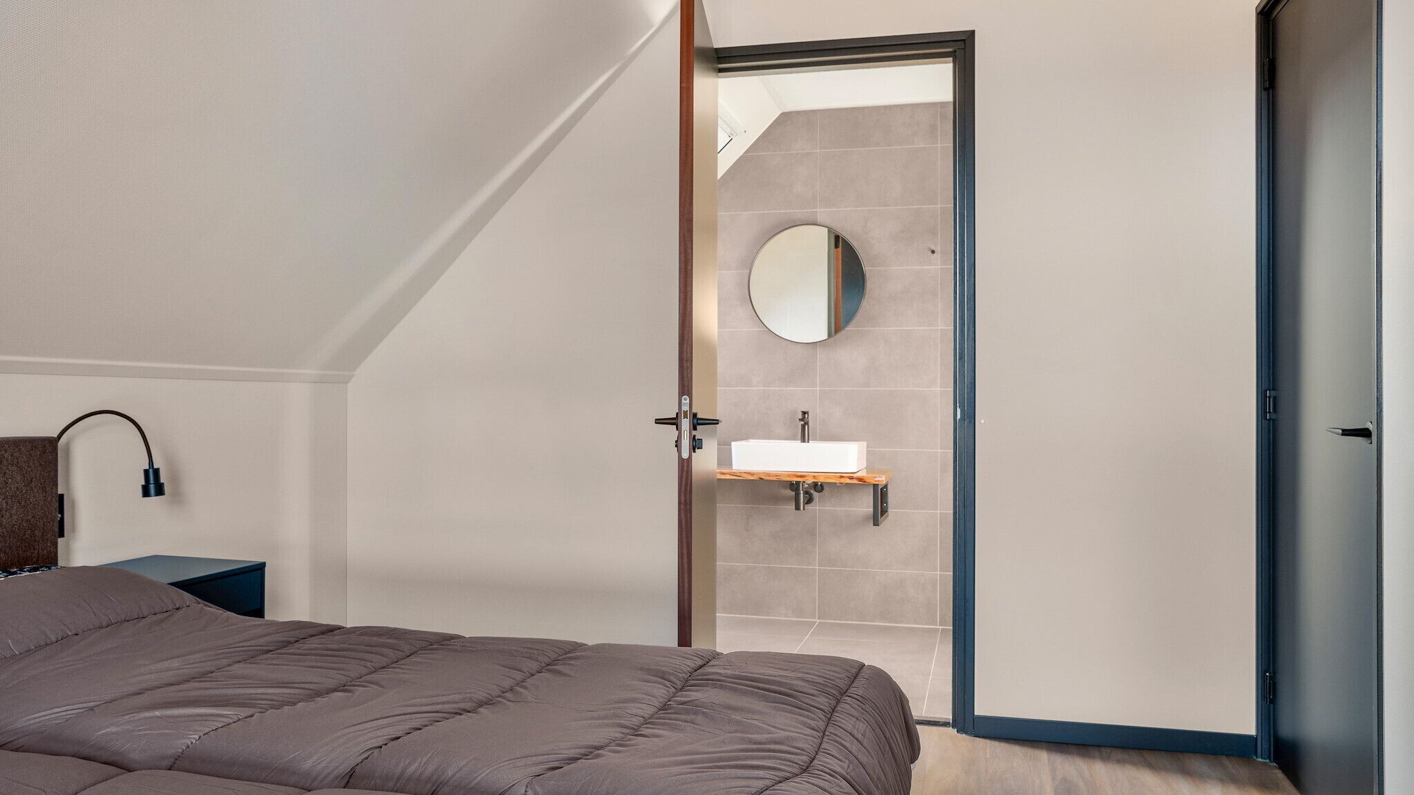 A bedroom with a bed, a bedside table, a mirror, and a bathroom with a sink and a towel rack.