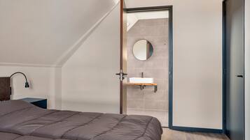 A bedroom with a bed, a bedside table, a mirror, and a bathroom with a sink and a towel rack.