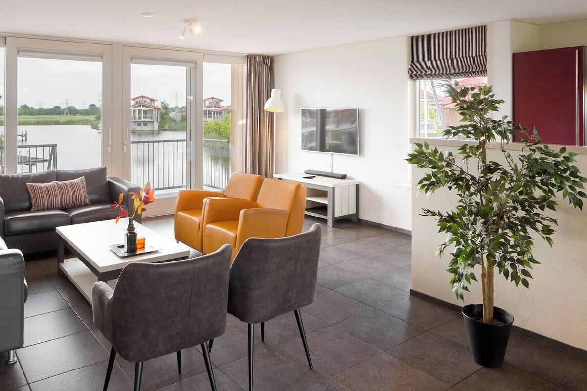 A modern living room with a sofa, armchairs, a coffee table, and a TV. There is a large window with a view of a body of water and buildings.