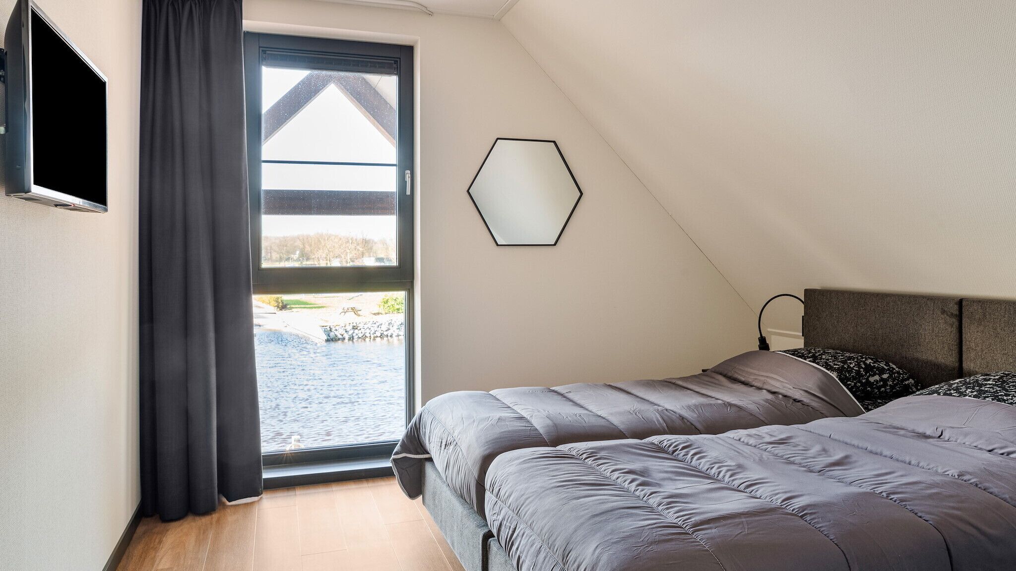 A bedroom with a large window, a hexagonal mirror, and a bed with grey bedding.