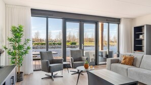 Una sala de estar moderna con un amplio ventanal corredera que da a un patio. Hay dos sillones, un sofá y una mesa de centro. Una planta en maceta se encuentra a la izquierda de la habitación.
