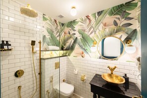 Une salle de bain avec des murs recouverts de carreaux blancs, une douche en verre, des toilettes, un lavabo avec un receveur doré et un miroir avec un cadre doré.