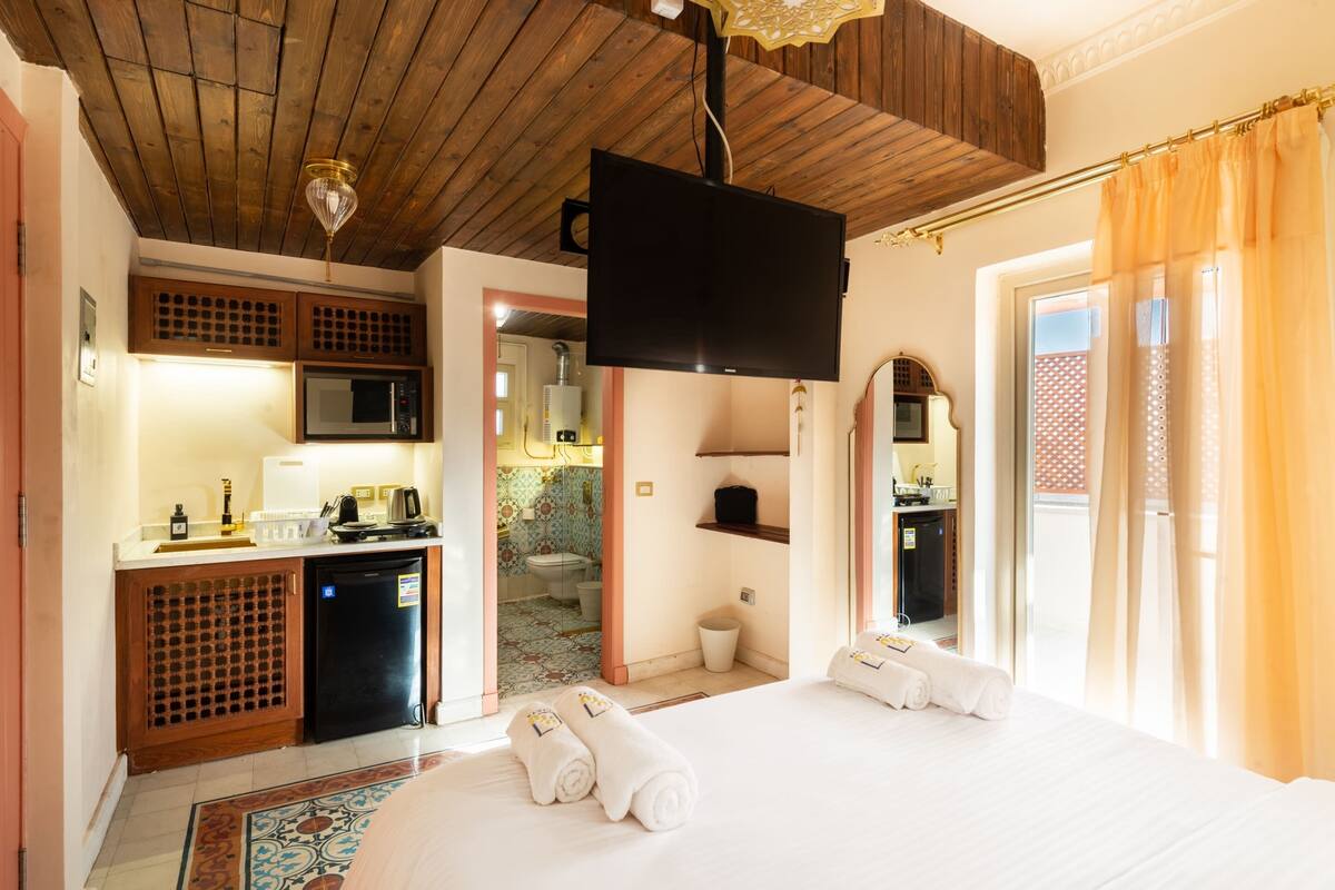 A hotel room with a bed, a flat-screen TV, a kitchenette, and a bathroom visible through an open door.