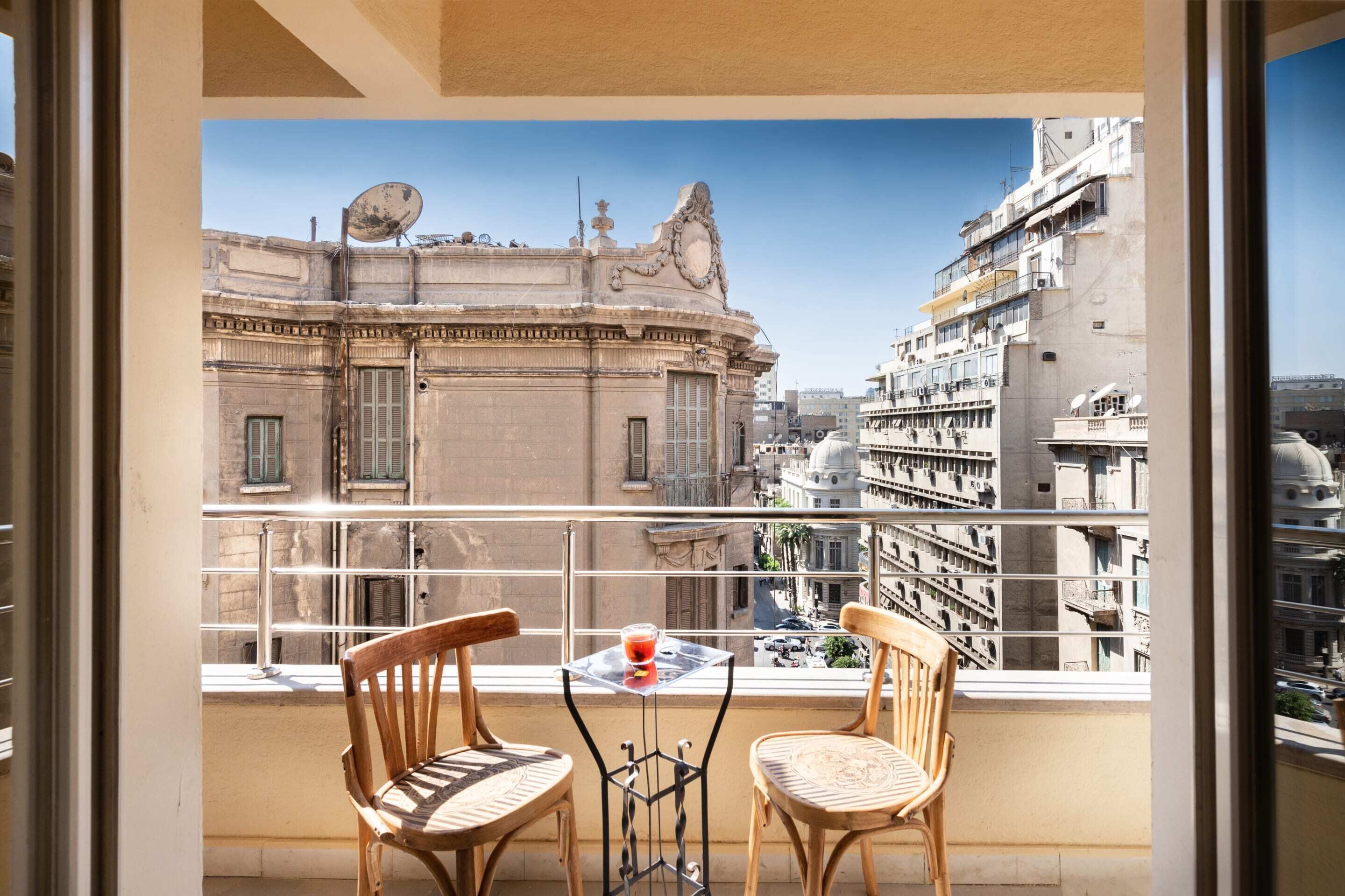 A balcony with two wooden chairs, a small table with a glass of red drink, and a view of historic buildings.
