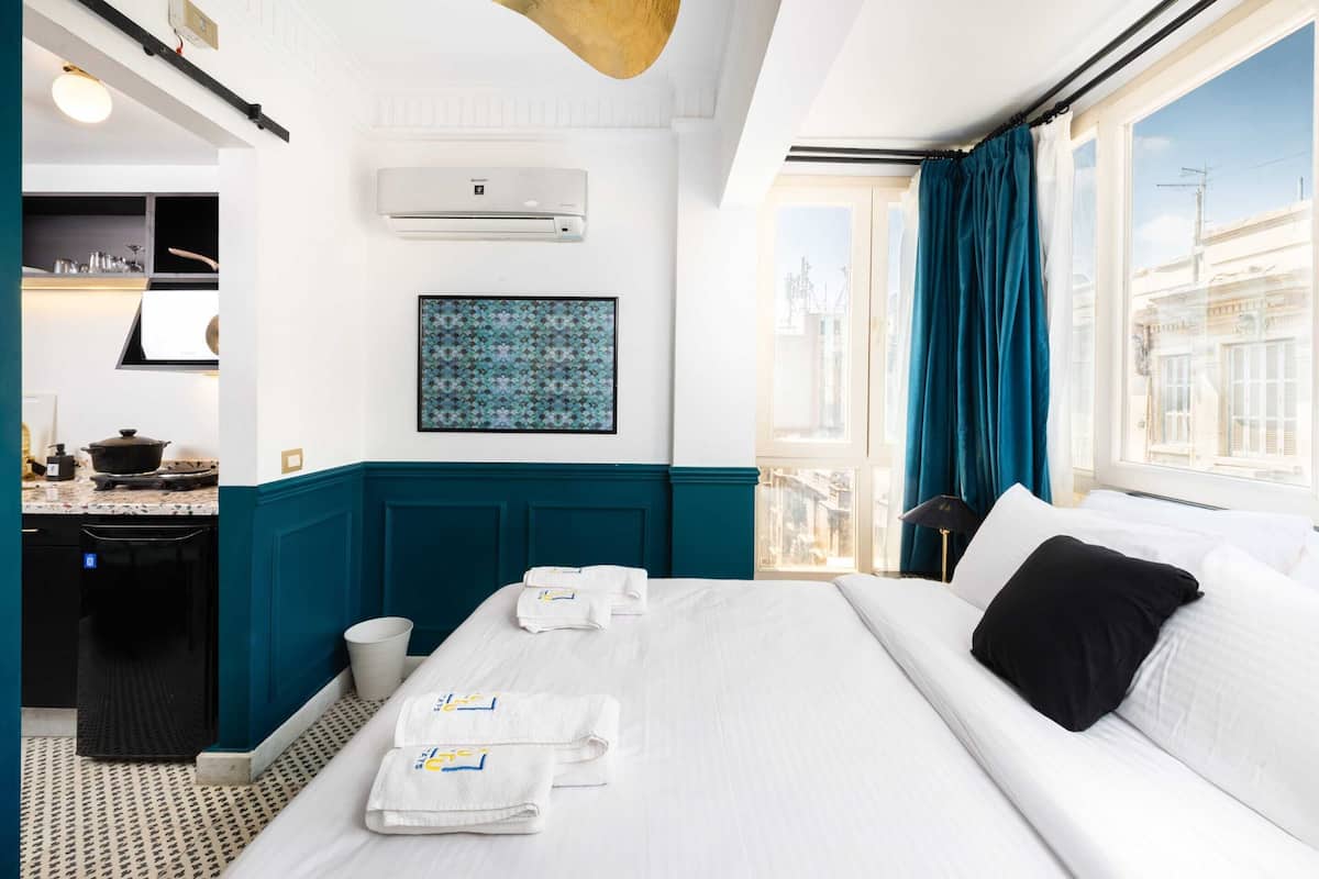 A hotel room with a bed, a desk, a kitchen area, and a view of the city.