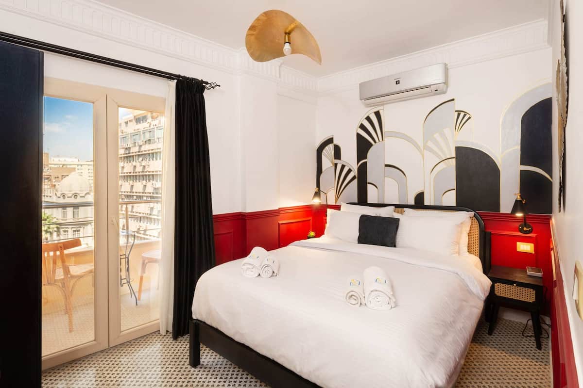 A hotel room with a large bed, a balcony with a view of buildings, a red accent wall, and a modern ceiling light.