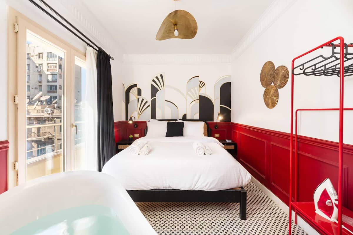 A hotel room with a large bed, a bathtub, a red metal rack, and a view of the city through a large window.